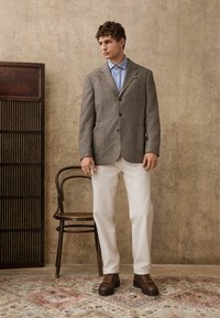 Gray patterned blazer with patch pockets, light blue shirt, white trousers, and brown lace-up boots on a textured beige background.