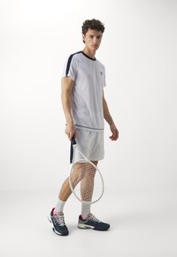 Hvit idretts skjorte med marineblå detaljer, hvite shortser, og mørkeblå tennissko med røde detaljer. Holder en tenniskølle.