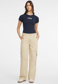 Jūrinis trumpomis rankovėmis marškinėliai su "GUESS JEANS" logotipu; smėlio spalvos kargo kelnės su šoninėmis kišenėmis ir tiesiomis kojomis; baltos sportbačiai su rudais padais.