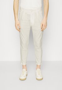 Muž v světlých béžových slim-fit kalhotách s kapsami a odpovídajících světlých béžových teniskách na jednobarevném pozadí.