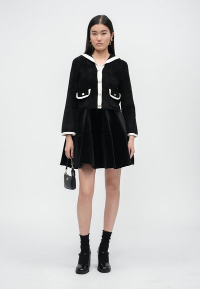 Schwarze Wildleder-Cropped-Jacke mit weißen Akzenten, Frontknöpfen und Taschen. Kombiniert mit einem schwarzen Samt-Hosenrock mit Schlag. Schwarze Schuhe und Socken. Kleine Handtasche.