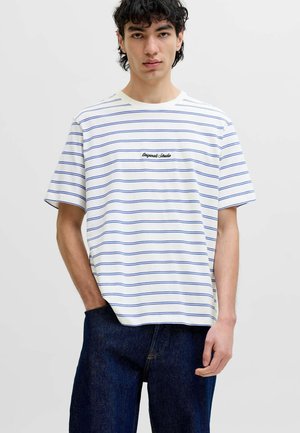 Mladý muž s kučeravými vlasmi, oblečený v biele tričko s krátkym rukávom s tenkými modrými horizontálnymi pásmi a tmavomodré džínsy.