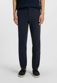 Granatowe spodnie o gładkiej fakturze, zwężanym kroju, z przednimi kieszeniami i zamkiem błyskawicznym, w zestawieniu z czarnymi butami wsuwanymi z kremową podeszwą.