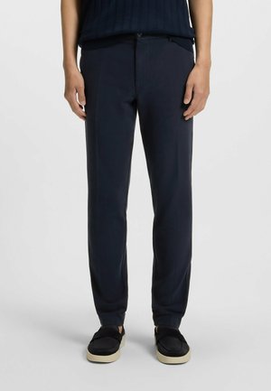 Navy blue trousers with a smooth texture, tapered fit, front pockets, and a zip fly, paired with black slip-on shoes featuring a cream sole.