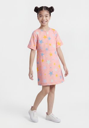 Mlada deklica z dvema figama, ki nosi roza obleko z večbarvnimi zvezdami in belimi supergami, nasmejana stoji na belem ozadju.