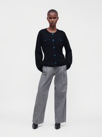 Femme debout les mains dans les poches, portant un cardigan noir boutonné avec des boutons bleus, un pantalon gris à jambes larges et des bottes noires.