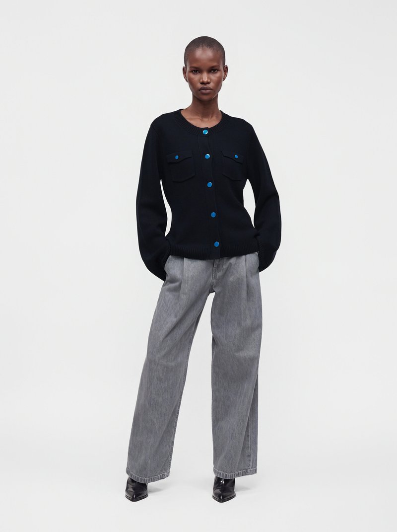 Femme debout les mains dans les poches, portant un cardigan noir boutonné avec des boutons bleus, un pantalon gris à jambes larges et des bottes noires.