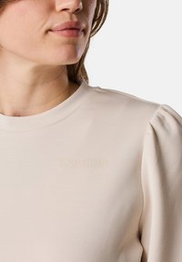 Top color crema con scollatura rotonda e maniche a sbuffo, con "SEASIDE" impressa in una tonalità chiara abbinata. Tessuto morbido e con texture.