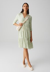 Robe vert clair avec un motif floral, col polo, manches trois-quarts et ourlet à volants. Portée avec des chaussures beiges et un petit sac.