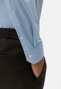Camicia azzurra chiara con polsini con bottoni, caratterizzata da un design angolato dei polsini e dettagli di cucitura minimi. Abbinata a pantaloni neri.