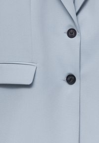 Blazer ajusté bleu clair avec deux boutons noirs et une poche à rabat sur le côté gauche.