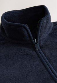 Gros plan d'une veste en polaire bleu marine avec un col montant et une fermeture éclair noire partiellement fermée.