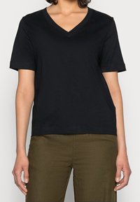 Schwarzes Baumwoll-T-Shirt mit V-Ausschnitt. Kurzarm und lockere Passform. Nahtdetails nahe dem Saum und den Ärmeln. Getragen mit olivgrünen Hosen.