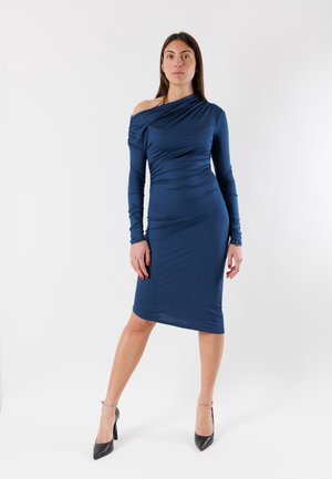 Donna in piedi che indossa un vestito aderente blu navy con una spalla scoperta e maniche lunghe, abbinato a tacchi neri, su uno sfondo bianco uniforme.