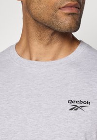 Pelēks kokvilnas T-krekls ar apaļu kaklu un īsām piedurknēm, uz kreisās krūts zonas izvietots melns Reebok logo. Gluda tekstūra.