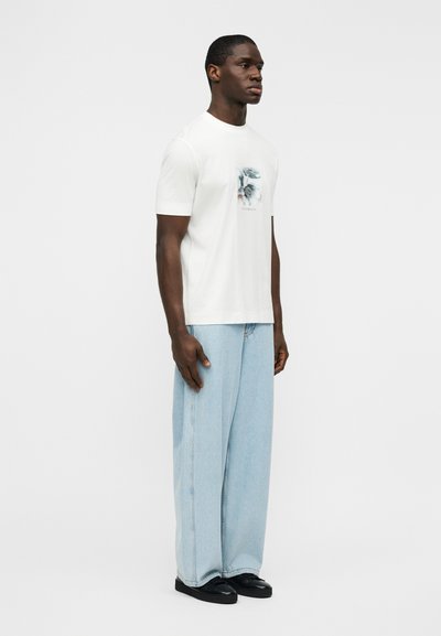 Λευκό βαμβακερό μπλουζάκι με πολύχρωμο γραφικό τύπωμα στο στήθος, συνδυασμένο με φαρδιά ανοιχτό μπλε τζιν παντελόνια και μαύρα αθλητικά παπούτσια.