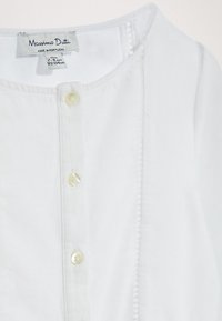 Valkoinen napitettava paita, joka on valmistettu pehmeästä kankaasta. Siinä on pyöreä kaula-aukko, etunapit pienillä pyöreillä napilla sekä hienovaraisia tekstuurityylejä.