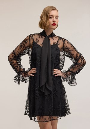 Abito nero con tessuto trasparente ricamato a fiori, maniche lunghe con polsini arricciati e dettaglio con sciarpa annodata al collo.