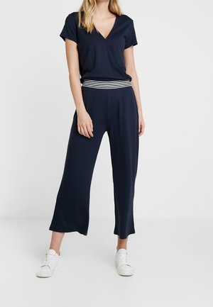 Kobieta ubrana w granatowy krótki top z dekoltem w serek i pasujące szerokie spodnie z paskiem w paski oraz białe tenisówki.