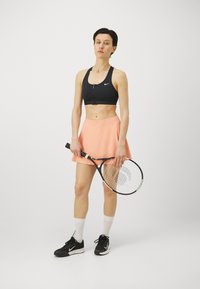 Zwarte sportbeha met ritssluiting aan de voorkant, perzikkleurige rok, zwarte tennissneakers met witte accenten, houdt een tennisracket vast. Glad stof, strakke pasvorm.