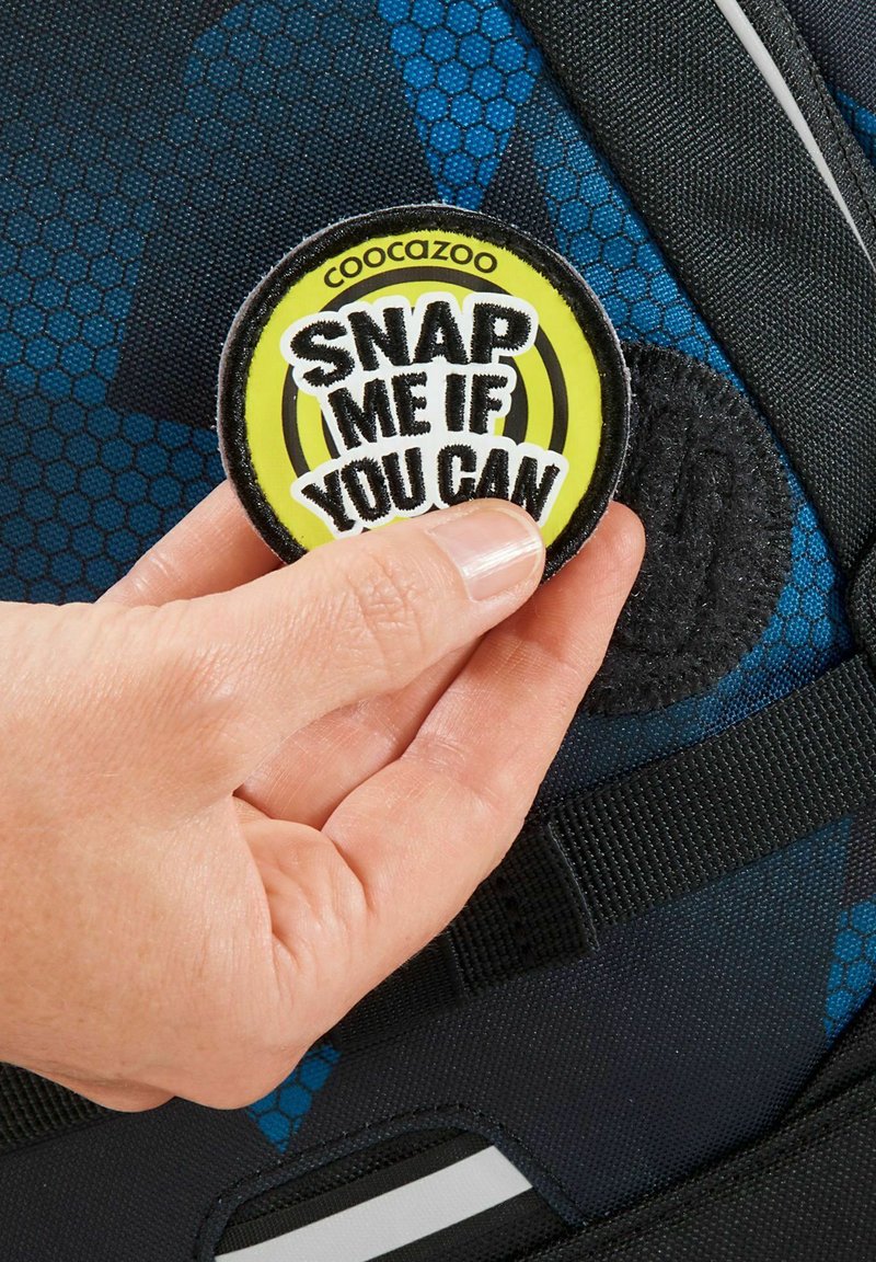 Runder Patch mit schwarzem Rand zeigt grünen und gelben Text "SNAP ME IF YOU CAN"; befestigt an einer gemusterten blau-schwarzen Stoffoberfläche.