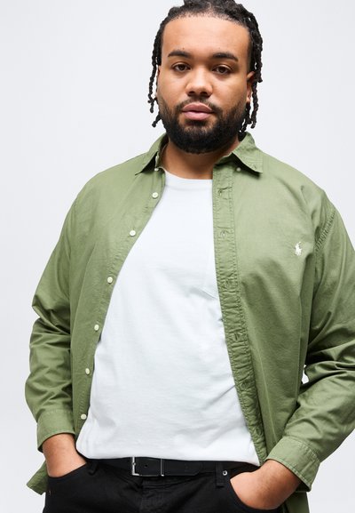 Άνδρας με πλεξούδες, φορώντας πουκάμισο σε χακί πράσινο με κουμπιά πάνω από λευκή μπλούζα, μαύρο παντελόνι, με τα χέρια στις τσέπες, ουδέτερο φόντο.