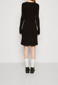 Abito lungo a maniche lunghe in maglia nera a coste con silhouette aderente, orlo sino al ginocchio e texture a coste. Indossato con calze bianche e scarpe con plateau.