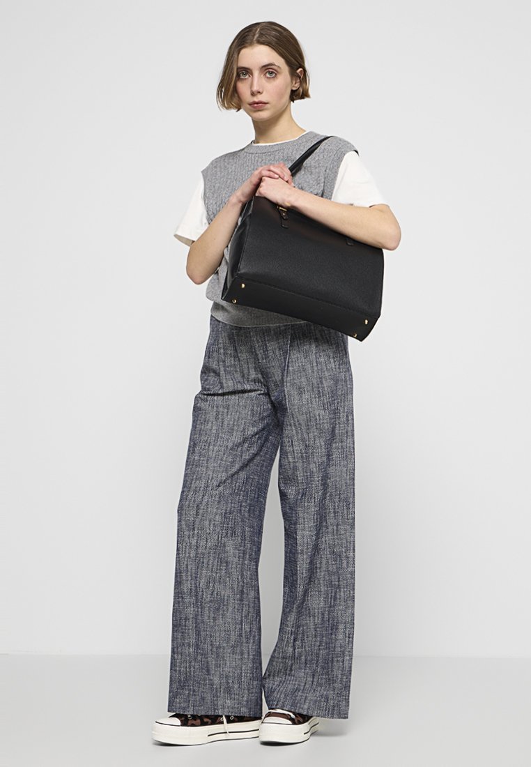 Borsa tote nera rettangolare con superficie testurizzata, dettagli in hardware dorato e una cerniera superiore. Il modello indossa un maglione grigio e pantaloni a gamba larga.