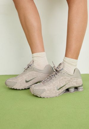 Ben iført cremefarvede sokker og lysegrå Nike-sneakers med fjedrede hæle står på grøn gulv.