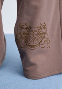 Brunt stof pryder et gyldent grafisk motiv af stiliserede katte langs siden, som fremviser en glat tekstur og et afslappet fit design.