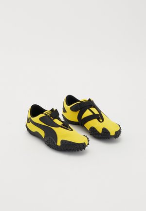 Paire de chaussures de sport jaunes et noires avec semelles texturées et sangles ajustables sur fond blanc uni.