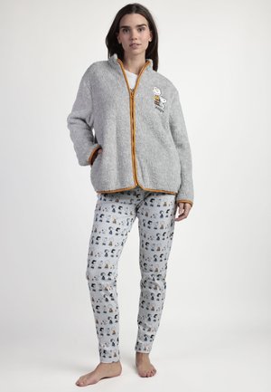 Mujer joven de pie descalza con chaqueta de forro polar gris con ribete naranja y bordado de Snoopy, y pantalones grises con estampados de personajes de dibujos animados.