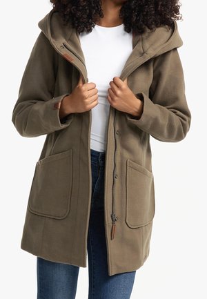 Femme portant un manteau à capuche vert olive avec de grandes poches devant, partiellement zippé sur une chemise blanche et un jean bleu.