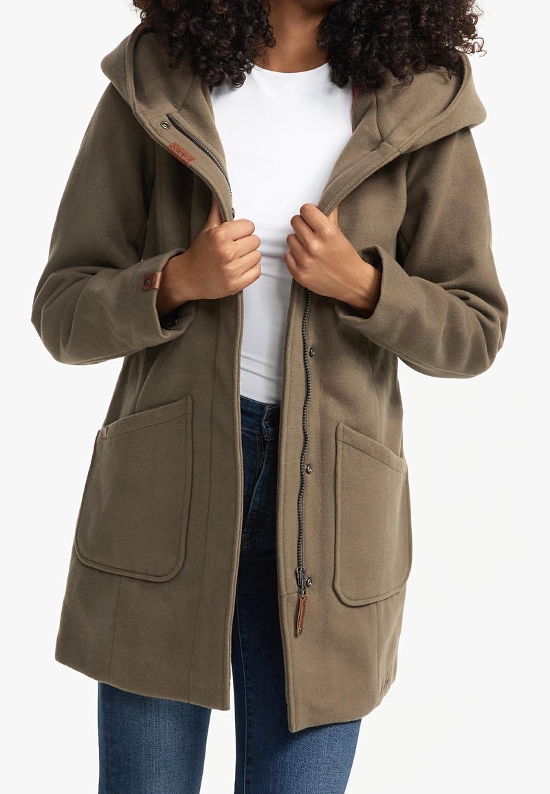 Femme portant un manteau à capuche vert olive avec de grandes poches devant, partiellement zippé sur une chemise blanche et un jean bleu.