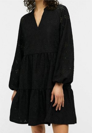 Woman wearing a black, long-sleeve, V-neck dress with eyelet lace fabric and a tiered skirt, standing against a plain background.