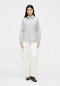 Γυναίκα που φοράει λευκό και μαύρο πουκάμισο με κάθετες ρίγες μακρυμάνικο, λευκό παντελόνι με φαρδύ πόδι και καφέ μποτάκια στον αστράγαλο, στέκεται μπροστά σε ένα απλό φόντο.