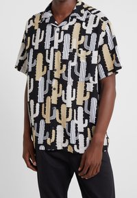 Chemise à manches courtes avec un motif de cactus en beige, noir et blanc. Fabriquée en tissu léger, avec un devant à boutons et un col.