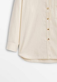 Camicia beige con bottoni, a maniche lunghe, caratterizzata da una texture liscia, orlo curvo e bottoni dorati lungo la parte centrale e la tasca.