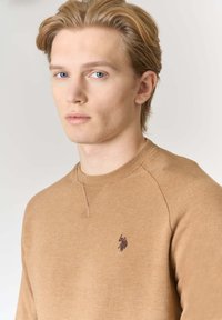 Felpa di cotone marrone chiaro con scollo rotondo, cuciture visibili e piccolo logo scuro sul petto a sinistra. Tessuto liscio.