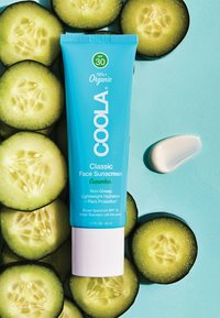 Coola Classic Gesichtssonnenschutz in einer blauen Tube mit weißem Deckel, umgeben von Gurkenscheiben auf einem hellblauen Hintergrund. Cremige Texturprobe vorhanden.