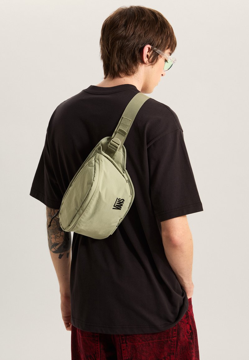 Sac bandoulière en tissu vert clair à texture lisse, avec un logo Vans, une sangle réglable et une forme compacte triangulaire.