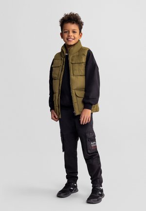 Chłopiec z kręconymi włosami uśmiechający się, ubrany w oliwkowo-zieloną kamizelkę puchową na czarnej bluzie oraz czarne spodnie cargo i trampki.