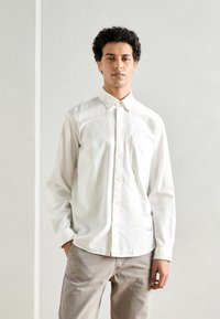 Camicia bianca in cotone con taschino sul petto e colletto classico, indossata con pantaloni beige chiaro. Tessuto morbido, vestibilità comoda.