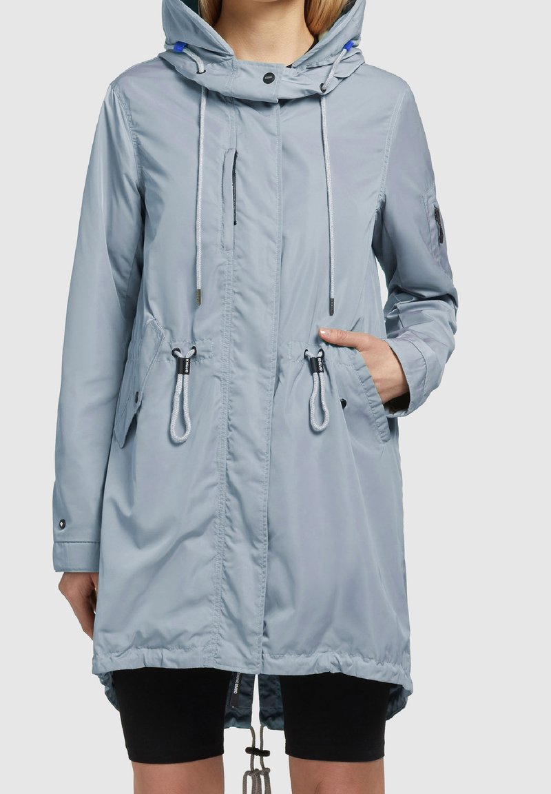 Femme portant un imperméable à capuche bleu clair avec poches avant et cordons de serrage, main dans la poche droite, short noir visible.