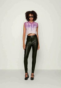 Jeune femme aux cheveux bouclés portant des lunettes de soleil noires, un pull sans manches violet, un pantalon en cuir noir et des talons noirs, debout.