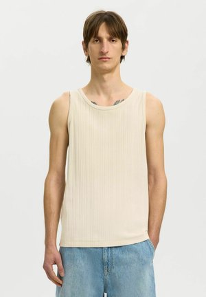 Homme portant un débardeur côtelé beige et un jean bleu clair, debout avec une main dans la poche devant un fond clair uni.