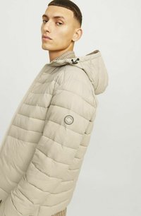 Veste matelassée beige avec capuche, panneaux cousus, fermeture éclair et un écusson avec logo sur la manche gauche. Texture lisse et légère.