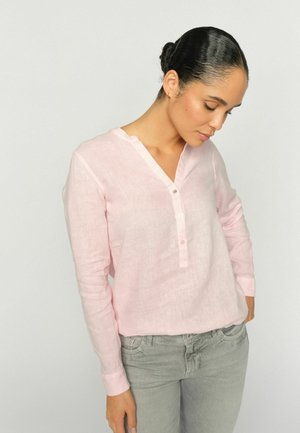 Woman with hair in bun wearing a light pink buttoned blouse and gray pants, looking down with hands near pockets against plain background.