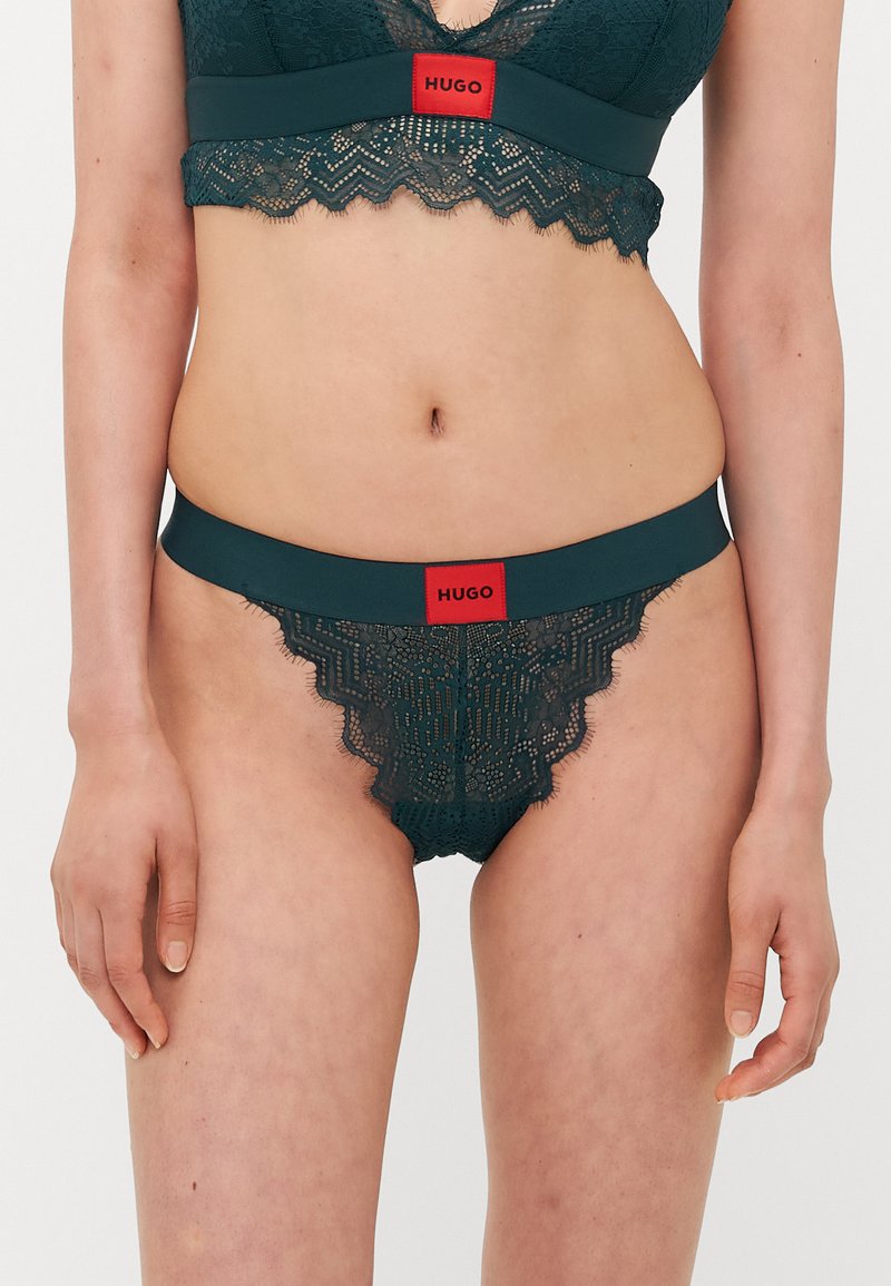 Mørkegrønn blonde-bralette og matchende truse med bølgete kanter. Har en solid kant med rød "HUGO"-logo i livet.