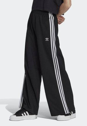Personne portant un pantalon large noir Adidas avec des bandes blanches sur les côtés et des baskets Adidas assorties noires et blanches, debout les mains derrière le dos.